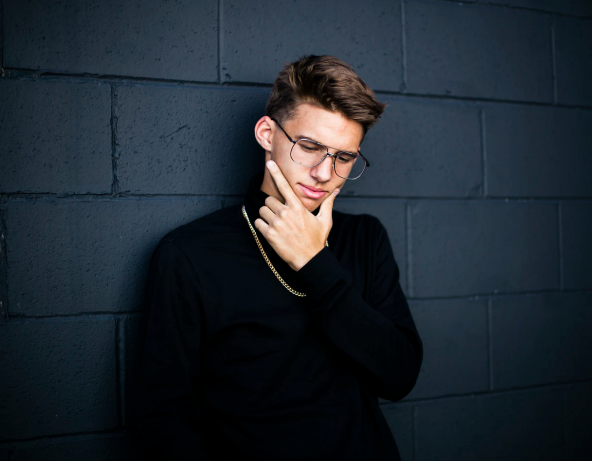 apply early , young male leaning against blue brick wall wearing glasses and rubbing his chin